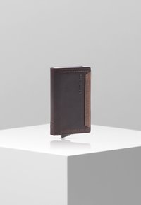 Portefeuille en cuir marron au design épuré, avec une face avant plus foncée et des accents plus clairs. Comprend des emplacements pour cartes et des détails en relief du logo.