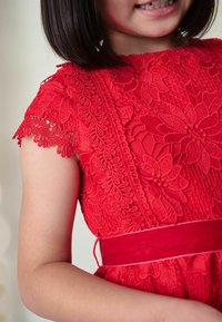 Czerwona koronkowa sukienka z kwiatowymi wzorami, krótki rękaw z misternymi detalami oraz pasującą czerwoną wstążką w talii.