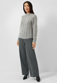 Grauer gestrickter Pullover mit einem strukturierten Muster, kombiniert mit weiten grauen Hosen und schwarzen Schuhen mit Absatz, vor einem neutralen Hintergrund.