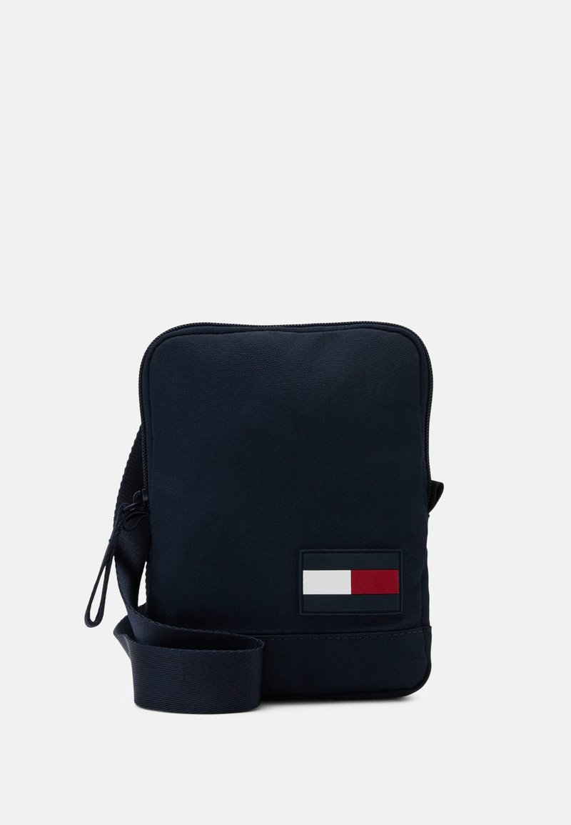 Kompaktní námořnická crossbody taška s nastavitelným popruhem a obdélníkovou nášivkou s logem v bílé, červené a černé barvě v dolním rohu na přední straně.