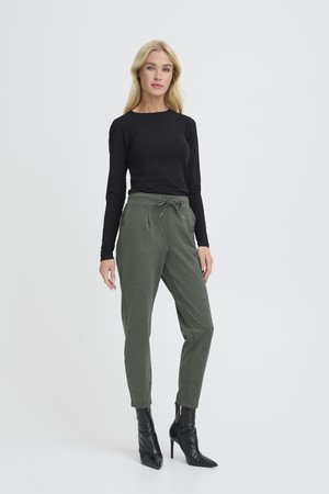 Schwarzes langärmliges Oberteil, kombiniert mit grünen Jogginghosen mit Kordelzug, seitlichen Taschen und schmal zulaufenden Beinen, getragen mit schwarzen Stiefeletten mit Absatz.
