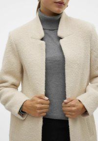 Abrigo de lana beige con un acabado texturizado, que presenta un cuello en pico y un frente abierto. Suéter de cuello alto gris visible debajo.
