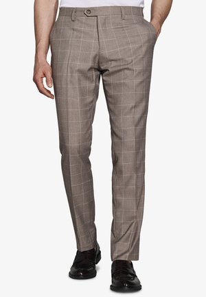 Mann trägt schlank geschnittene braune karierte Hose mit Taschen, kombiniert mit schwarzen Lederschuhen, steht vor einfachem Hintergrund.