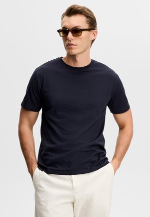 Muž v tmavomodrom tričku a bielych nohaviciach s rukami v vreckách, pozerá sa do strany, nosí hnedé obdĺžnikové slnečné okuliare.