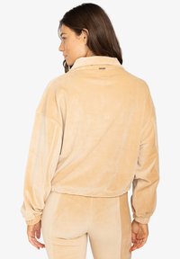 Giacca pullover in corduroy beige con colletto alto, spalle scese e texture rigata. Presenta polsini elasticizzati e un dettaglio logo discreto.