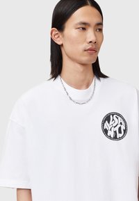 Mlada osoba s dugom crnom kosom nosi bijelu oversized majicu s okruglim crnim AllSaints logotipom na prsima i srebrnim lančićem.
