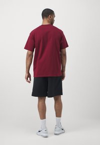 T-shirt de algodão bordô com mangas curtas, combinado com calções pretos. O modelo usa sapatilhas brancas e meias de cano médio, visto de costas.