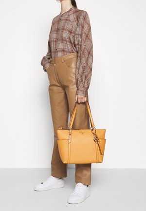 Persoon draagt een bruin geruit overhemd met knopen, beige leren broek, witte sneakers, en houdt een grote gele handtas met dubbele bovenhandvatten vast.