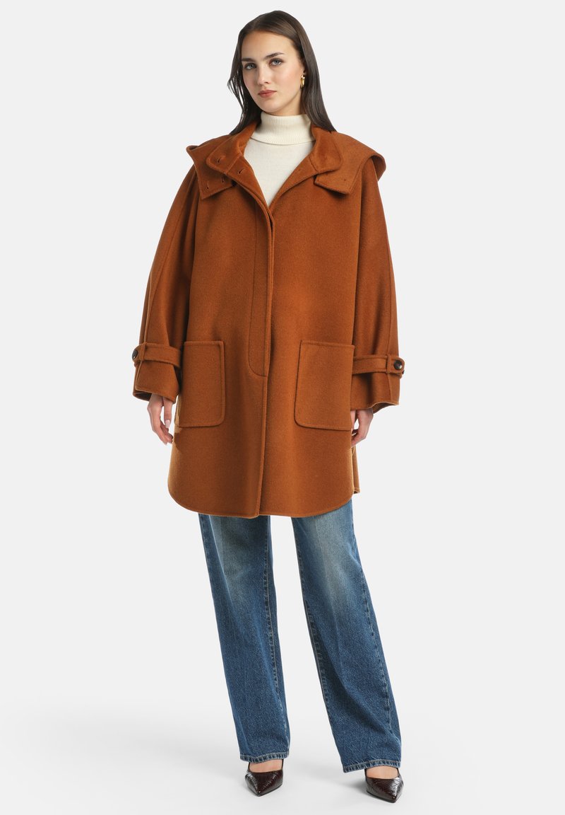 Cappotto di lana marrone con cappuccio, grandi tasche anteriori e maniche con bottoni; indossato sopra un dolcevita crema e jeans a gamba larga blu.