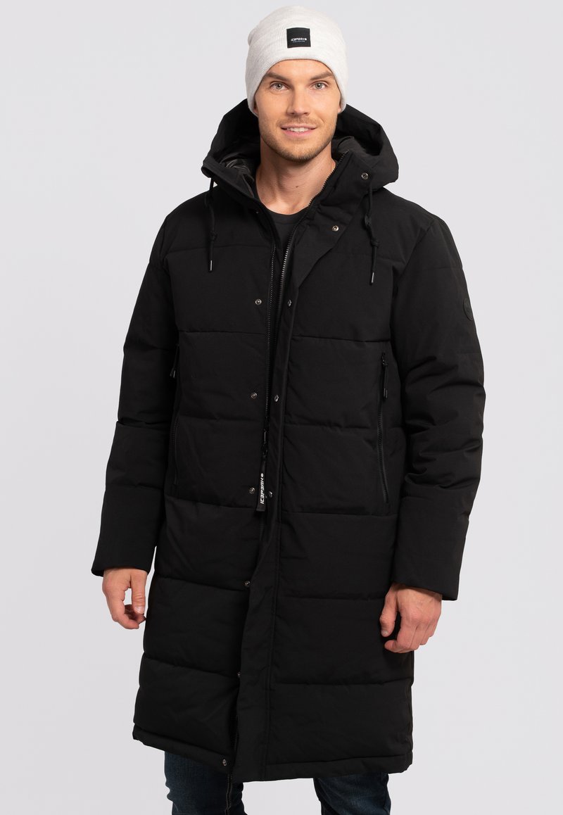 Abrigo negro acolchado con capucha, que presenta un cierre de cremallera y botones a presión, dos bolsillos laterales y un diseño acolchado; llevado con un gorro ligero.