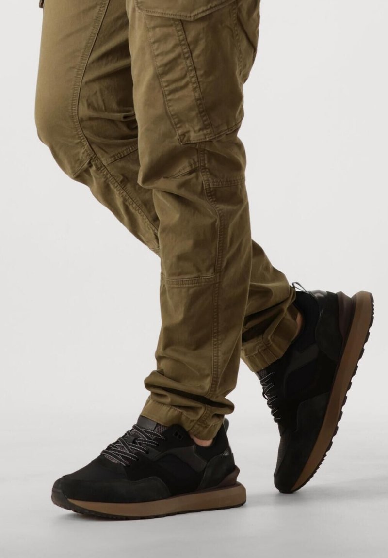 Zwarte sportieve schoenen van suede en mesh materialen, met grijze veters en een bruine rubberen zool. Gecombineerd met olijfgroene cargobroek.