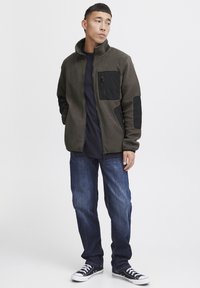 Olijfgroene fleece zip-up jack met zwarte accenten, voorzien van een borstzak en gestructureerde mouwen, gecombineerd met blauwe jeans en zwarte sneakers.