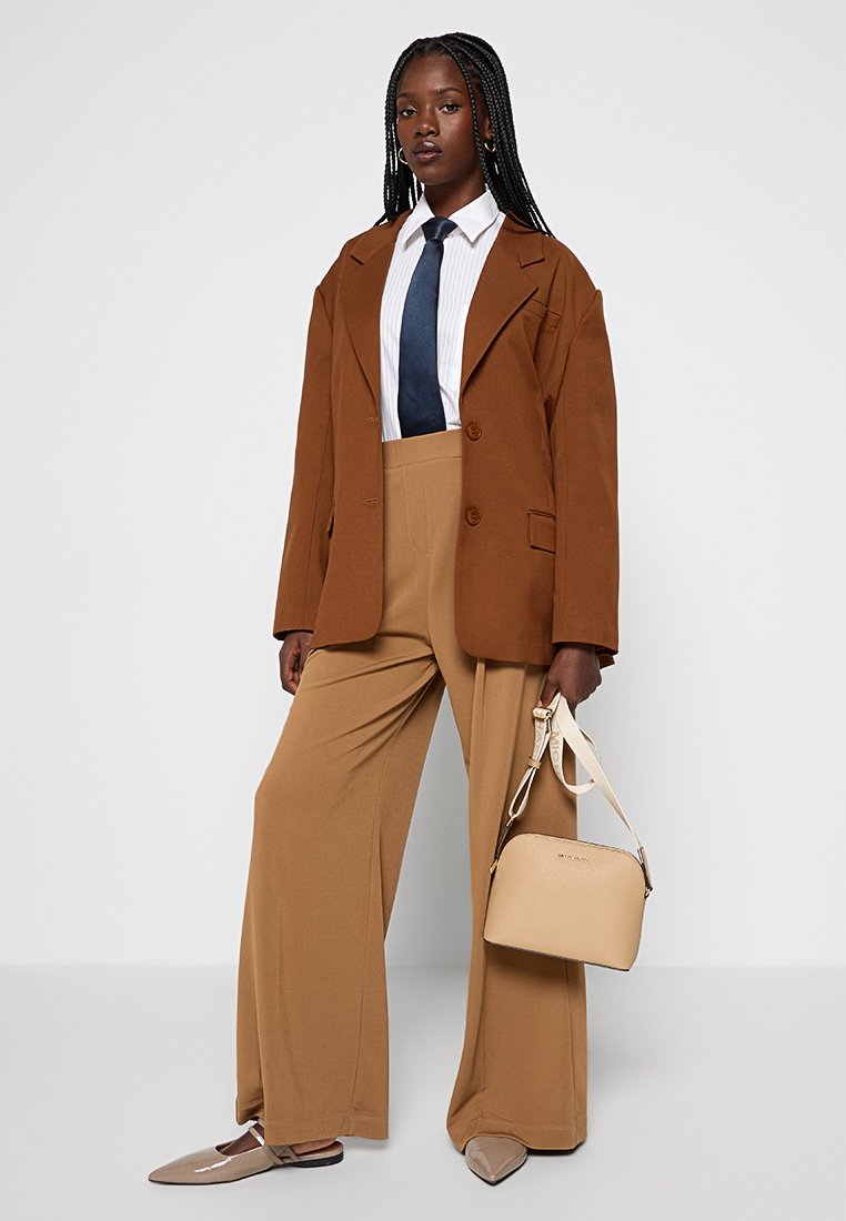 Blazer brun surdimensionné, chemise rayée blanche, cravate bleue marine, pantalon beige à jambe large et un sac à main neutre avec une courte anse. Textures douces, coupe ajustée.