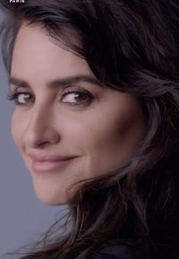 Eine Nahaufnahme des Gesichts einer Frau mit langen, dunklen, welligen Haaren. Sie hat braune Augen, dezente Konturierung und hellen Lippenstift vor einem grauen Hintergrund.