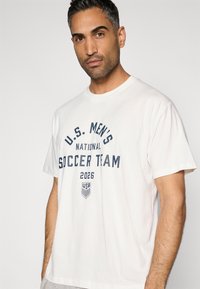 Camisola de algodão branca com texto azul que diz "EQUIPA NACIONAL DE FUTEBOL MASCULINO DOS EUA 2026" acima de um pequeno logótipo de futebol dos EUA. Mangas curtas.