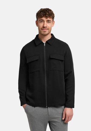 Man wearing black zip-up jacket with two chest pockets and gray pants, standing against plain white background.