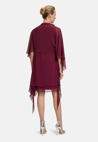 Burgunderfarbenes Kleid mit geschichteten, transparenten Kapuzenärmeln und asymmetrischem Saum, kombiniert mit silbernen High Heels. Glatter, fließender Stoff.