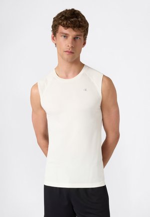 Jonge man met kort krullend haar, gekleed in een wit mouwloos sportshirt en zwarte korte broek, staand tegen een effen achtergrond.