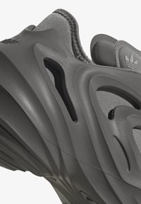 Grijze sport schoen met een gestructureerde bovenkant, kenmerkend golvend ontwerp, uitsparingen voor ventilatie, en contrasterende gladde en matte afwerkingen.