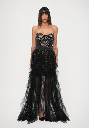 Donna in un abito senza spalline di tulle nero e trasparente, con un corpetto di paillettes e una gonna a strati, in piedi di fronte a uno sfondo bianco semplice.