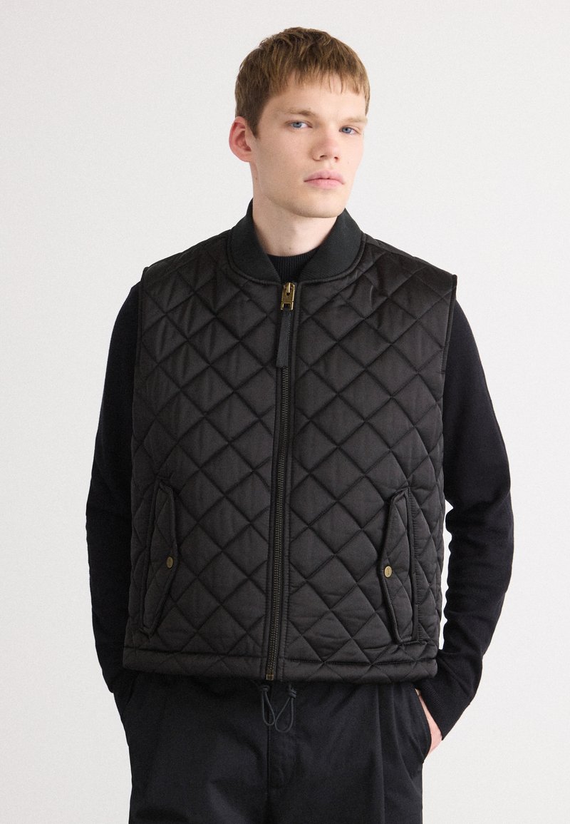 Black quilted vest with a high collar, front zip closure, side pockets, and a textured pattern. Soft fabric with a smooth finish.