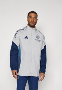 Lys grå sportsjakke med marineblå ermer. Har Adidas-logo, Arsenal-emblemet og glidelås foran. Glatt tekstur, lett design.