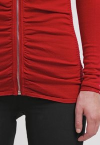 Röd långärmad topp med dragkedja fram och rynkad tyg, parat med svarta byxor, som visar en persons torso och högra hand.