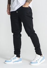 Jeans negros de corte slim con cierre de botón, que presentan una textura suave y un diseño ajustado, combinados con zapatillas deportivas blancas y azules.