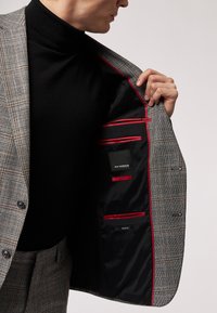 Giacca da abito a quadri grigi, con fodera nera e dettagli rossi e diversi taschini interni. Chiusura frontale con bottoni e vestibilità sartoriale.