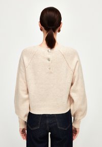 Femme aux cheveux bruns attachés en queue de cheval basse, portant un pull beige avec trois boutons dans le dos et un jean bleu foncé, vue de dos.