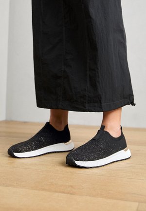 Fødder iført sorte slip-on sneakers med hvide såler og glitrende detaljer, stående på trægulv, delvist dækket af sort lang nederdel.
