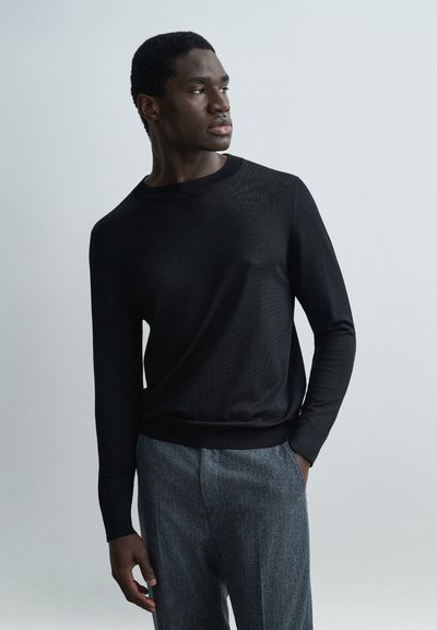 Homme en chemise noire à manches longues et pantalon gris, debout avec une main dans la poche, regardant sur le côté sur un fond clair uni.
