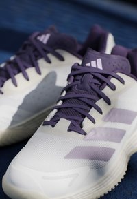 Valge ja lilla Adidas spordijalatsite paar paeladega sinisel pinnal, näidates ees- ja külgede lähivaate detaile.