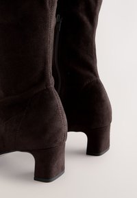 Stivaletti alla caviglia in suede marrone con tacco basso a blocco, dotati di cerniera laterale e finitura testurizzata. Il design enfatizza una silhouette liscia e aderente.