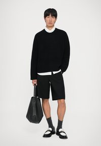 Maglione nero a coste, camicia con colletto bianco, pantaloni corti neri, calzini grigi, scarpe nere con dettagli bianchi, mentre tiene una grande borsa a tracolla nera.