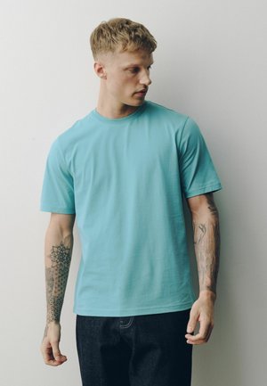 Fiatal férfi rövid szőke hajjal, kék pólót és fekete nadrágot visel, mindkét alkarján geometriai és természet témájú tetoválásokat mutatva.