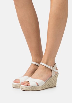 Valkoiset wedge-sandaalit ristikkäisillä nauhoilla ja nilkkahihnalla. Jute-tyylinen kanta lisää tekstuuria ja korkeutta. Silkkinen viimeistely täydentää muotoilua.