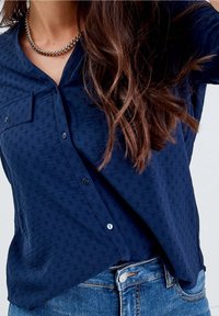Femme portant une chemise boutonnée texturée bleu marine avec une poche poitrine, un collier en chaîne dorée et un jean en denim bleu.