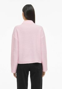 Jasnoróżowy sweter z prążkowanym kołnierzem i mankietami, o luźnym kroju i fakturowanym wzorze dzianiny. Noszony z czarnymi spodniami.