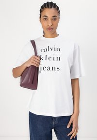 Balta medvilnės marškinėliai su juodu spaudu "calvin klein jeans". Modelis laiko mažą bordo odinį krepšį. Žemiau matyti džinsai.
