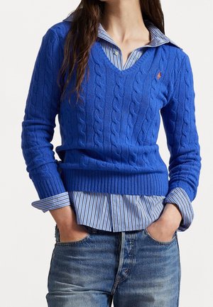 Personne portant un pull bleu à col en V en maille torsadée sur une chemise à col rayée bleue rentrée dans un jean bleu délavé, les mains dans les poches.