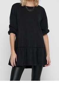 Kobieta w luźnej, czarnej bluzce z długim rękawem, z detalem marszczonego dołu i podwiniętymi mankietami, w połączeniu z błyszczącymi, czarnymi legginsami i wielowarstwowym złotym naszyjnikiem.