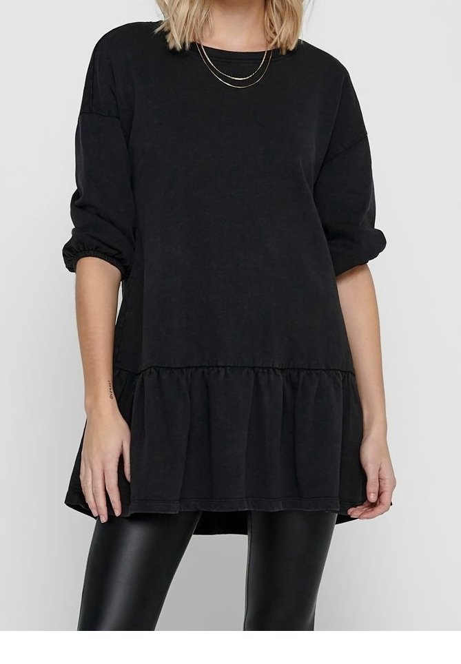 Kobieta w luźnej, czarnej bluzce z długim rękawem, z detalem marszczonego dołu i podwiniętymi mankietami, w połączeniu z błyszczącymi, czarnymi legginsami i wielowarstwowym złotym naszyjnikiem.