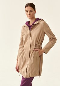 Giacca leggera beige con vita cinchata, chiusura con zip e cappuccio. Presenta tasche laterali e un tessuto morbido e texturizzato.