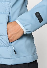 Lichtblauwe jas met gestikte textuur, ritssluitingszak aan de zijkant en zwart logo patch op de mouw. Zachte stof met elastische manchetten.