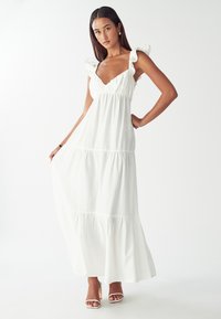 Nainen, joka on pukeutunut hihattomaan valkoiseen maksimekkoon, jossa on röyhelöolkaimet, pidellen hieman hameen helmaa, seisoo yksinkertaista valkoista taustaa vasten.