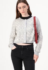 Lys grå kort cardigan med et teksturert blomsterdesign, har to knapper og ribbede mansjetter, kombinert med svarte høytlivsjeans.