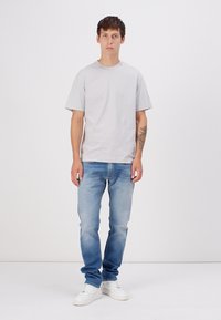 Gray cotton t-shirt with short sleeves, paired with light blue jeans. Model wears white sneakers, standing against a plain white background.