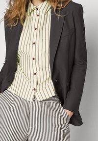 Mustat bleiseri vaaleanvihreän raidallisen paidan päällä, jossa on viininpunaiset napit, yhdistettynä musta-valkoisiin raidallisiin housuihin. Tekstuuritiedot näkyvissä.