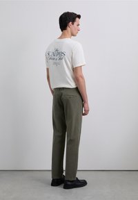 Man in wit T-shirt met de tekst "The Scalpers Taste & Talk", olijfgroene broek en zwarte schoenen, staand met zijn gezicht naar een effen grijze muur.
